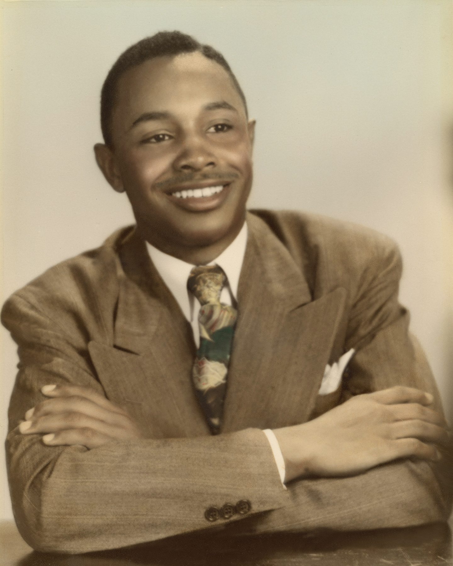 Hand-tinted studio portrait of Hezekiah Moore in 1947, smiling, arms folded, in a light suit and patterned tie.
